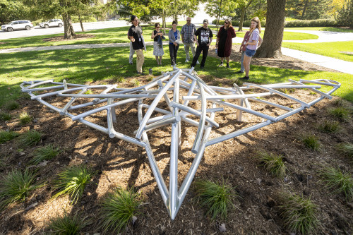 A tour group stops to discuss a metal sculpture shaped like a bird in flight. It's made of large metal poles that resemble sticks, criss crossing to make the outline of the bird's body. Its shape references the effigy mounds also found on campus.