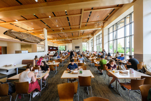A wide view of a student dining hall with large windows and a wooden ceiling. A birchbark canoe hangs from the rafters.