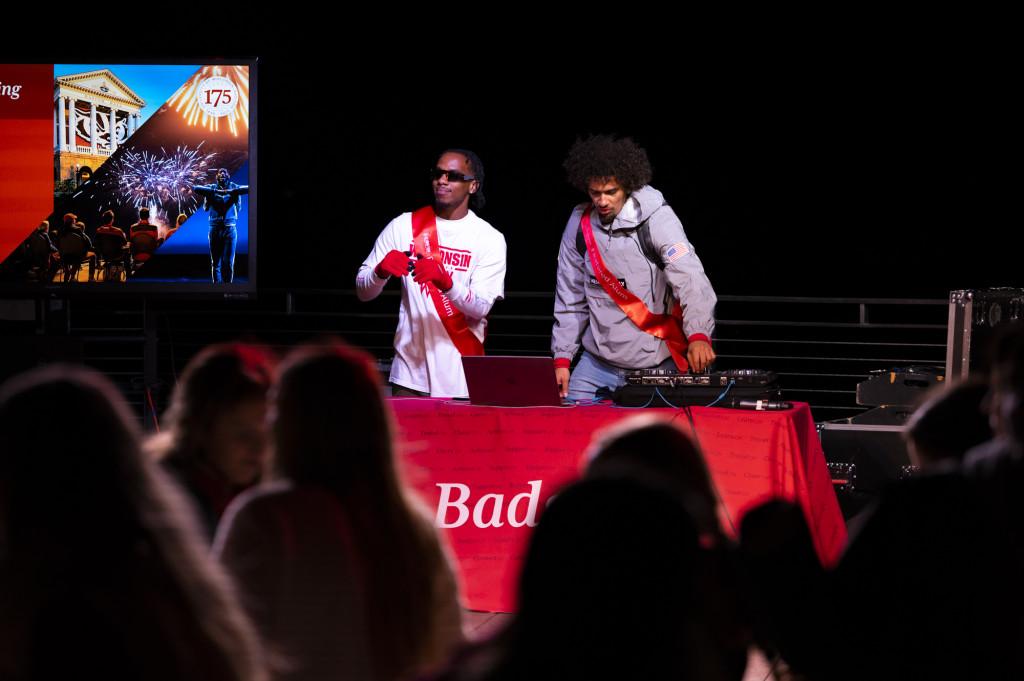 In a photo taken at night, two men stand on stage at a table covered in a red cloth. They're working a DJ setup and interacting with the crowd. Both men are wearing red sashes that say Featured Alum in white text.