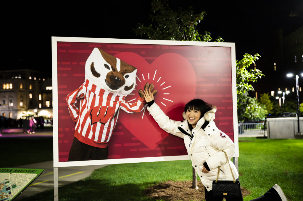 In a photo taken at night, a woman in a white puffy jacket smiles to the camera as she high fives with a poster of Bucky Badger gesturing a high five in return.