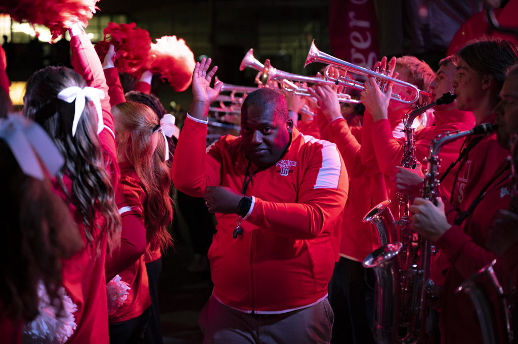 Standing amid trumpeters and members of the Sprit Squad, Corey Pompey holds his arms to make an L sign as he conducts the brass section of the UW Marching Band.