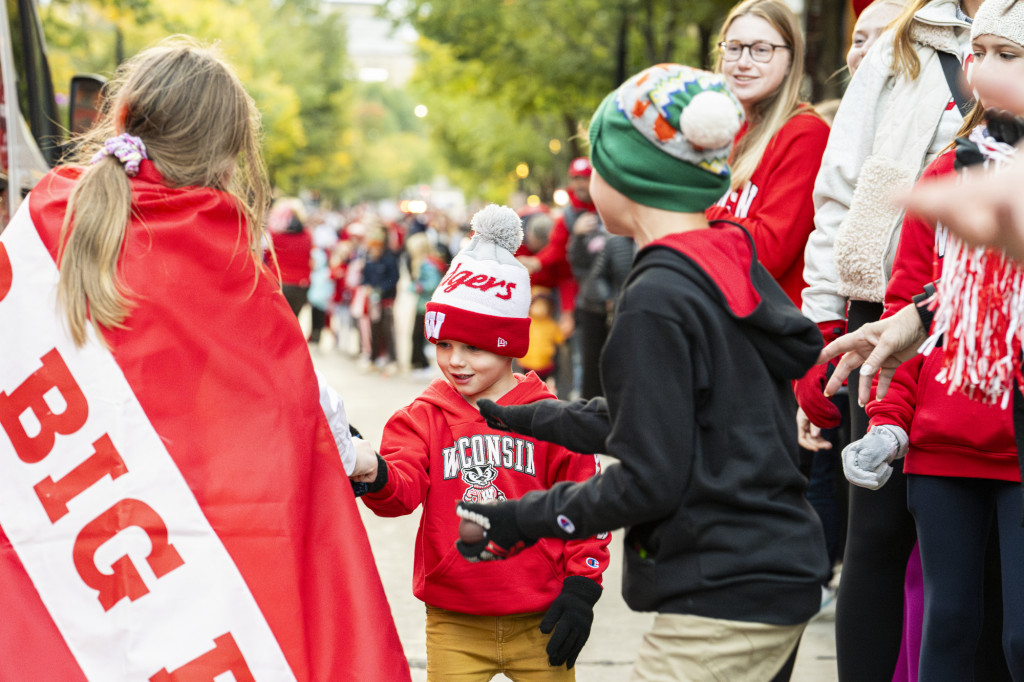 Facing away from the camera, a woman in a red and white cape hands candy to two young children as adults look on.