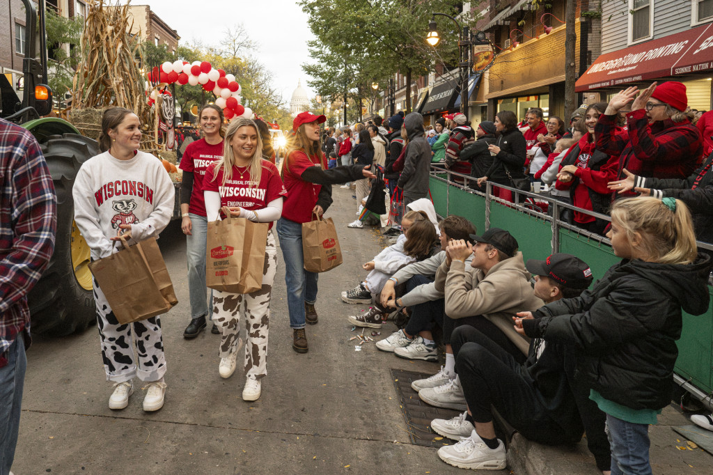 Dressed in Wisconsin T-shirts and sweatshirts, Four women holding paper grocery bags throw candy to crowds gathered along state street. The women are marching in the Homecoming parade. Behind them is an agriculture-themed float being pulled by a John Deere tractor.