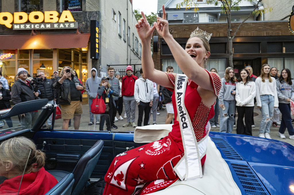 A woman wearing a tiara, a white Miss America sash and a red dress makes a W sign with her hands as she cheers along with the crowd. She's seated on top of a convertible as it rolls down State Street.