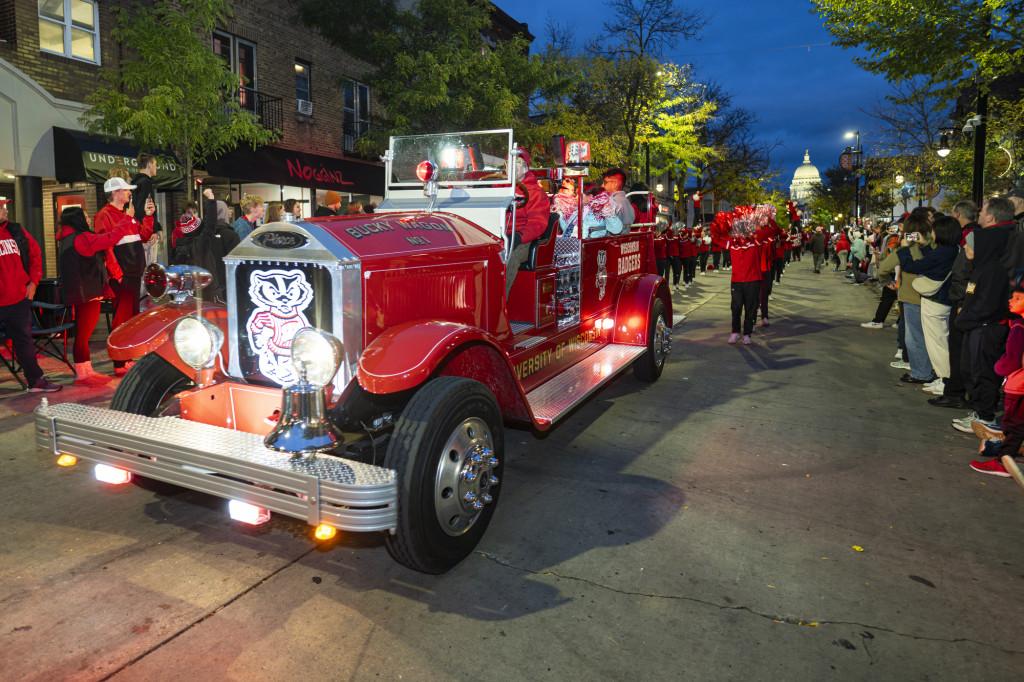 In a photo taken at night, an antique fire engine pained red and decorated with decals of Bucky Badger drives down State Street, leading the UW Spirit Squad. The capitol dome shines in the background.