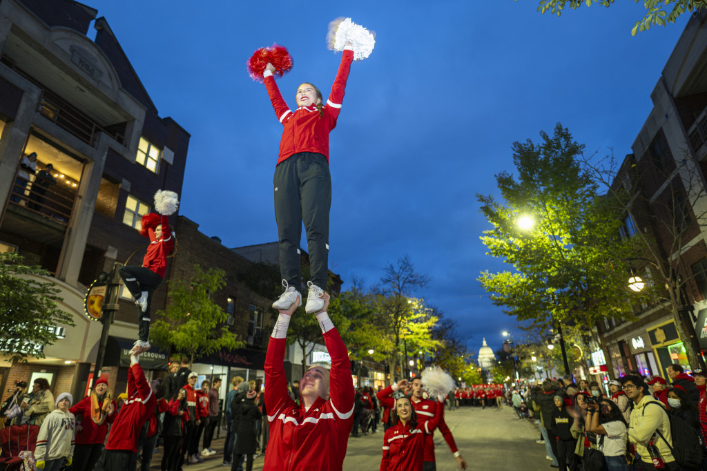 A man holds a woman aloft, supporting her feet as she stands above him with her hands in the air waving pompoms.