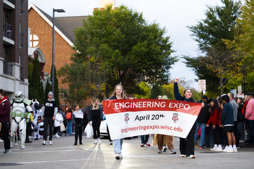 Among the paraders, two women carry a banner that says Engineering Expo April 20th engineeringexpo.wisc.edu.