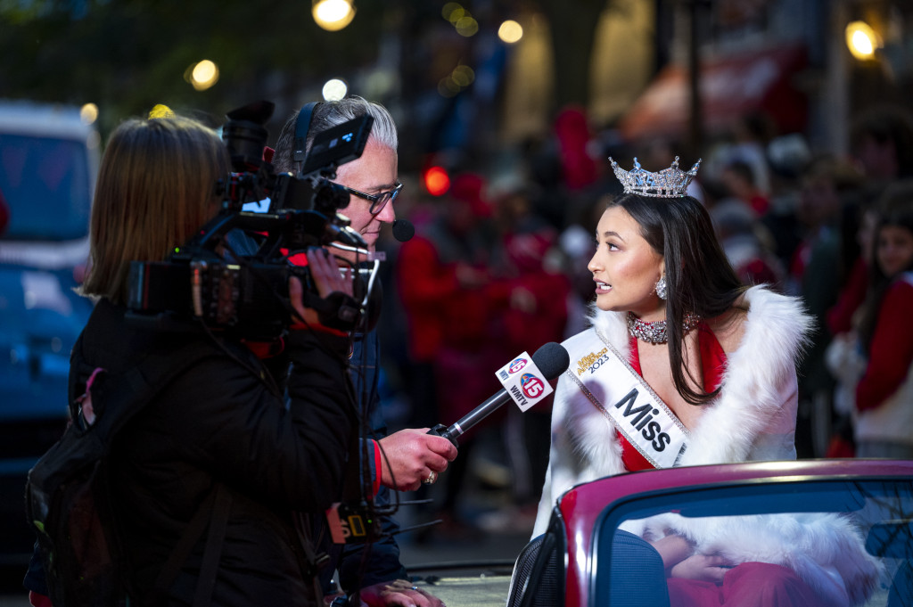 In a photo taken at night, a TV camera light illuminates Miss Wisconsin as she speaks into a microphone being held by a news reporter. She is wearing a tiara, a white sash and a red dress. She is seated in an open convertible.