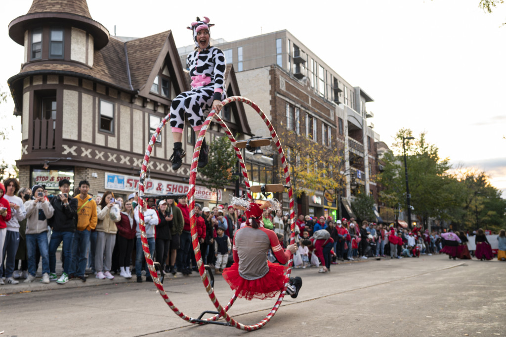 Two acrobats, one dressed as a black and white cow, the other in a red tutu, ride a large wheel down the parade route as the crowd cheers them on.