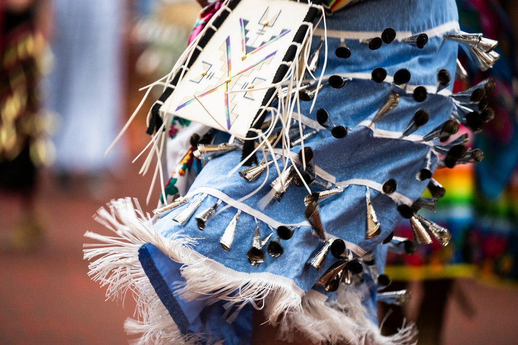 A close shot of Indigenous regalia in motion. The blue hem is adorned with shining metal and tassels.