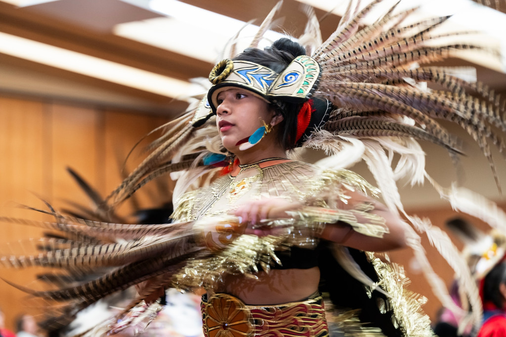 A woman wearing a head piece adorned with long, pointed pheasant feathers, spins as she dances.