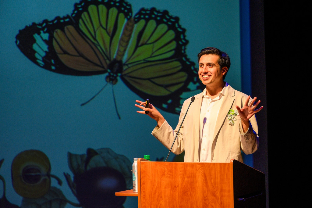 A speaker stands at a podium with images of butterflies appearing on the screen in the background.