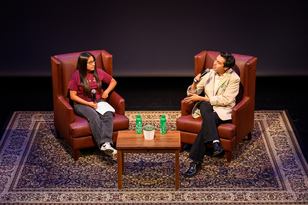 Two people sit in chairs on a stage engaging in a conversation.