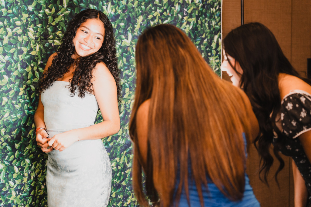 A woman poses in front of a leaf-printed backdrop while two friends take a photo.