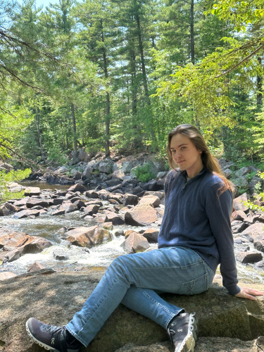 A portrait photo of Nick Kaska. Nick is sitting on a large rock in a river bed among tall, green trees.