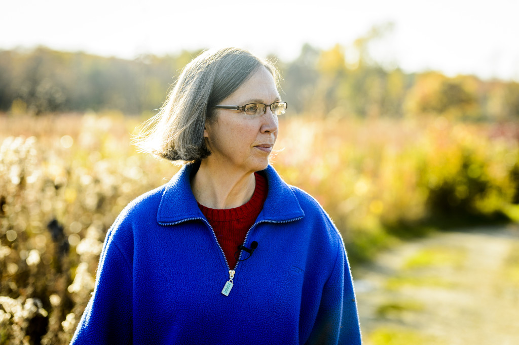 A woman wearing a blue fleece with a lapel mic clipped to her collar looks to the left as she walks down a path through a prairie.
