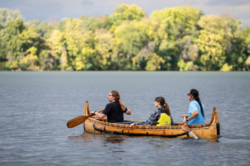 Two people paddle a birchbark canoe in Lake Mendota while a UW student rides in the center of the craft.