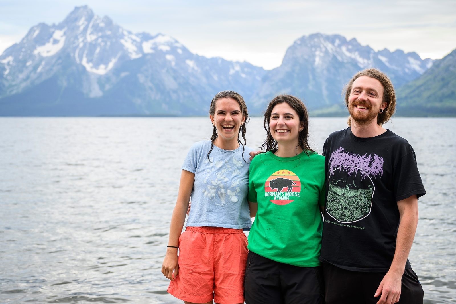 Lucy McGuire, Arielle Link and Timon Keller stand shoulder to shoulder smiling and laughing in front of the calm blue waters of Jackson Lake. The Teton Mountains rise up dramatically behind them speckled by patches of snow.