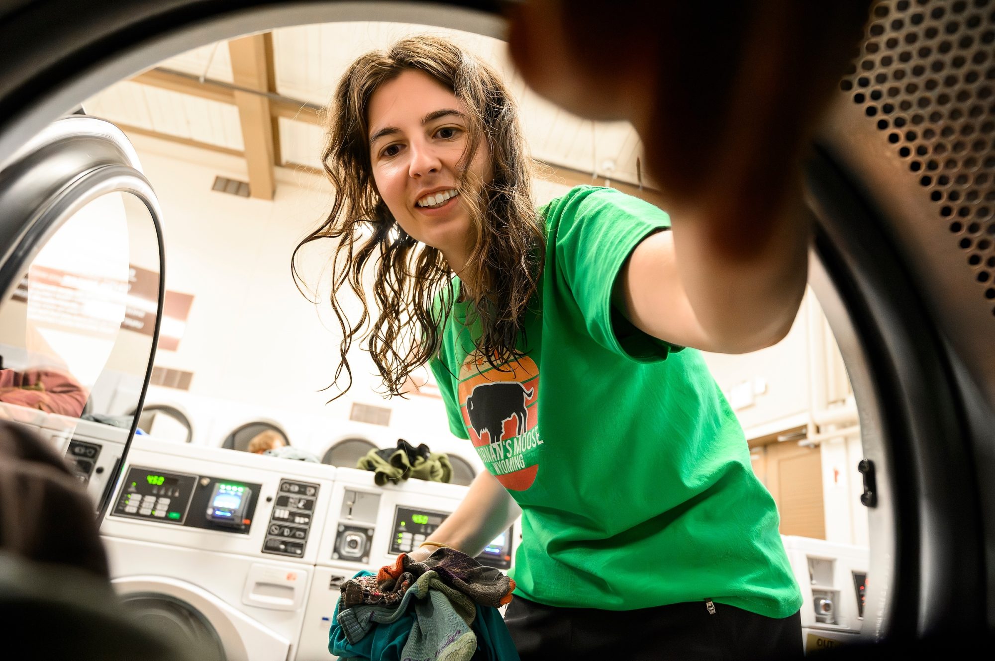 Looking out from inside a dryer, Arielle Link is smiling and holds a handful of colorful hiking socks while she moves her laundry over from a washing machine.