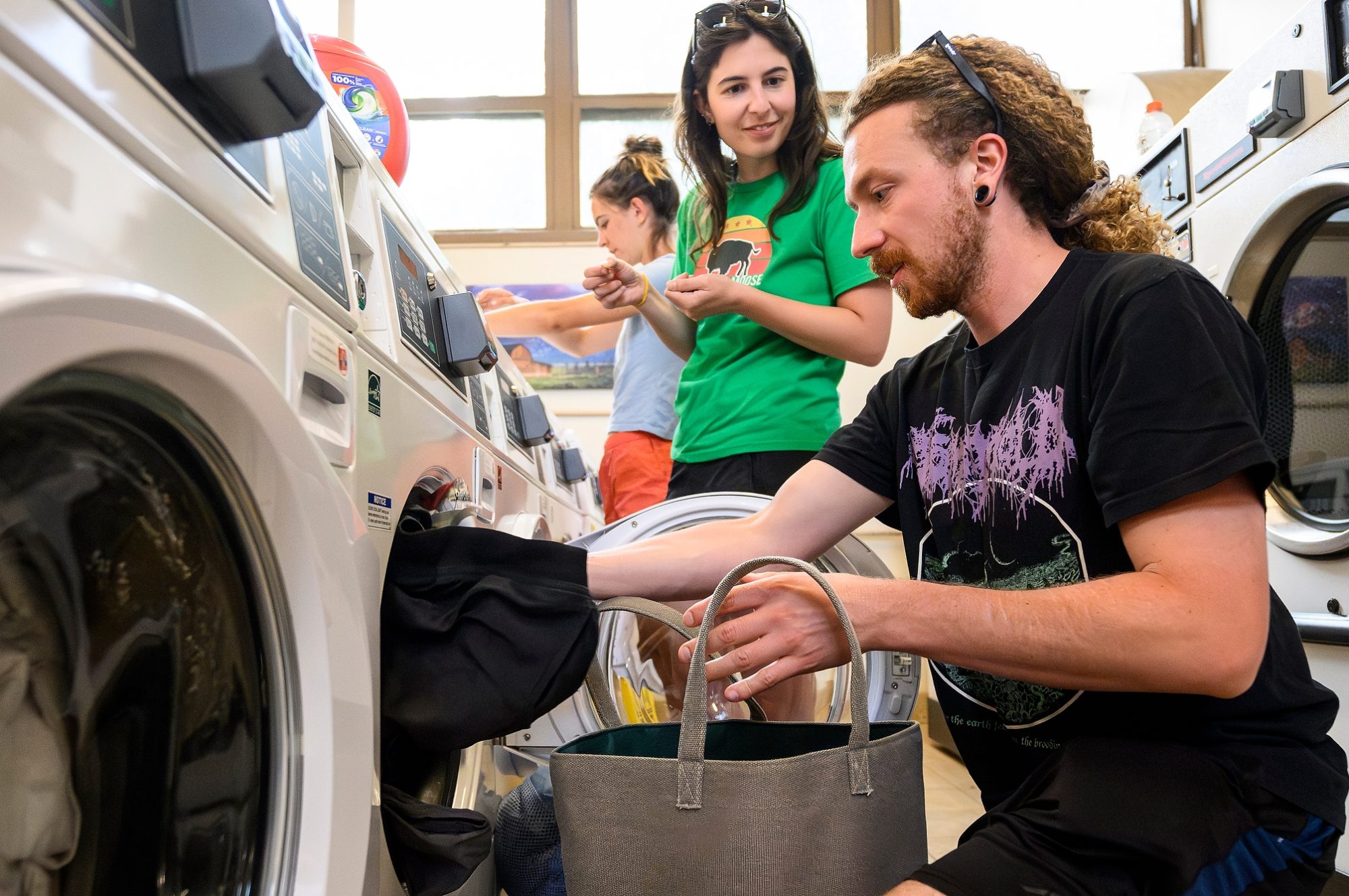 Timon Keller squats down in the foreground to load his laundry into a washing machine at a laundromat lined with washer and dryers. Behind him, Arielle Link and Lucy McGuire also prepare to put their own laundry into machines.