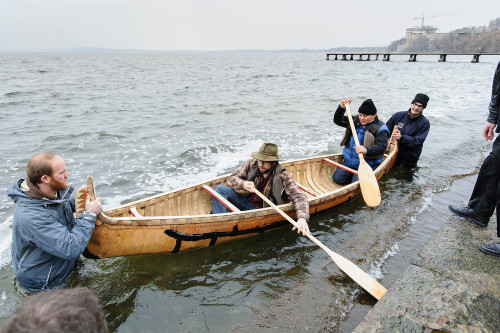 Two men in waders stand in waist-deep water holding the ends of a birchbark canoe steady whole two men with paddles sit in the canoe, ready to push off from shore.