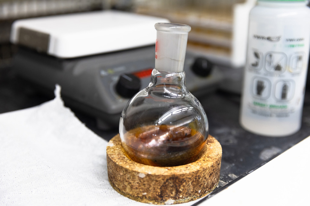 On a laboratory bench, a glass beaker with a round bottom holds a brown liquid and sits on a wooden coaster.