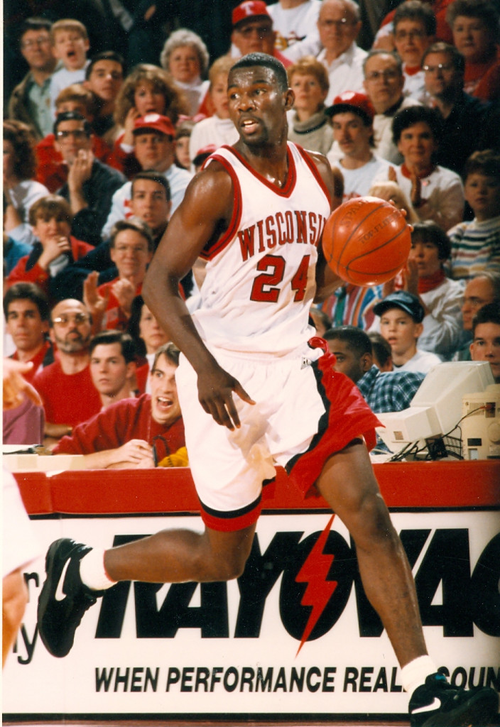 A man in a Wisconsin Badgers jersey dribbles the basketball as the crowd watches him.
