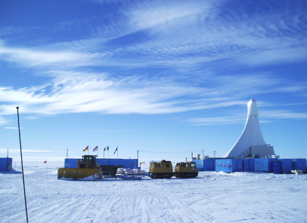 A photo of a base camp set up on an Antarctic ice sheet. A cluster of blue shipping containers surround a tall teepee-like structure that towers over the containers.