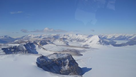 An aerial view through an airplane window of a frozen Antarctic landscape. Ice and snow covers a jagged mountain range.
