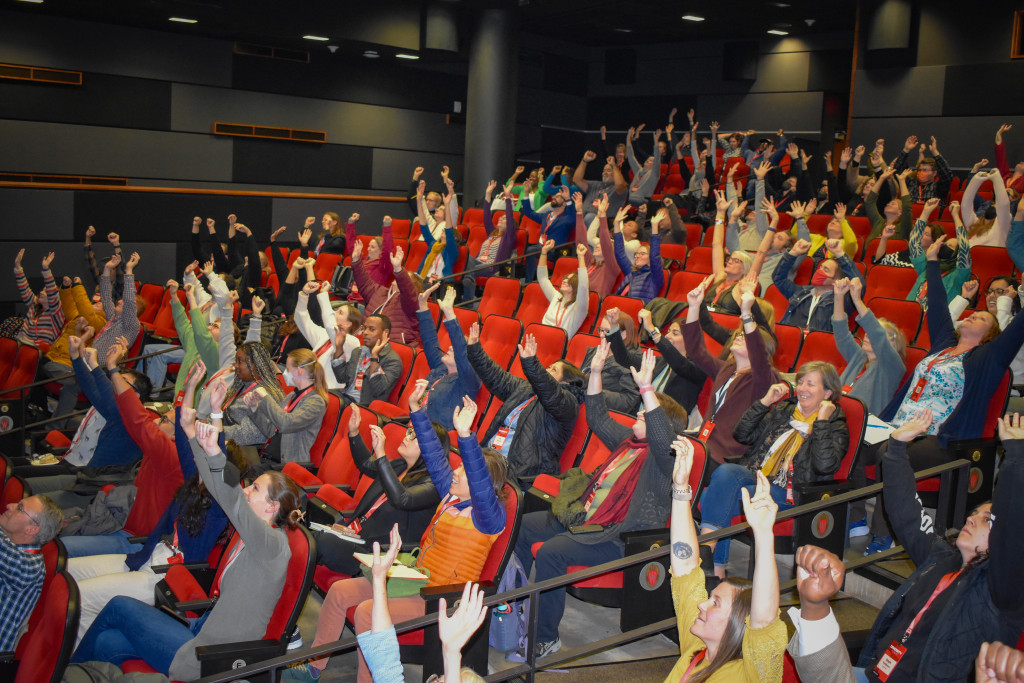 People in an auditorium raise their hands and smile.