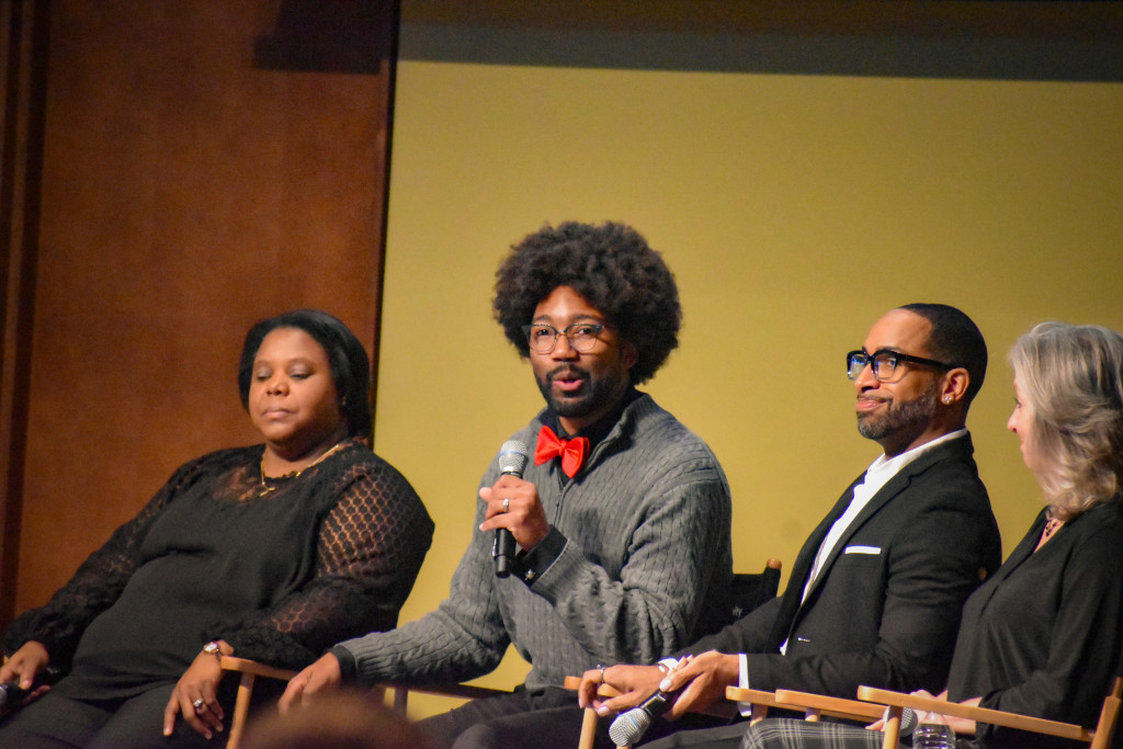 Several people sit in chairs, one talks into a microphone.