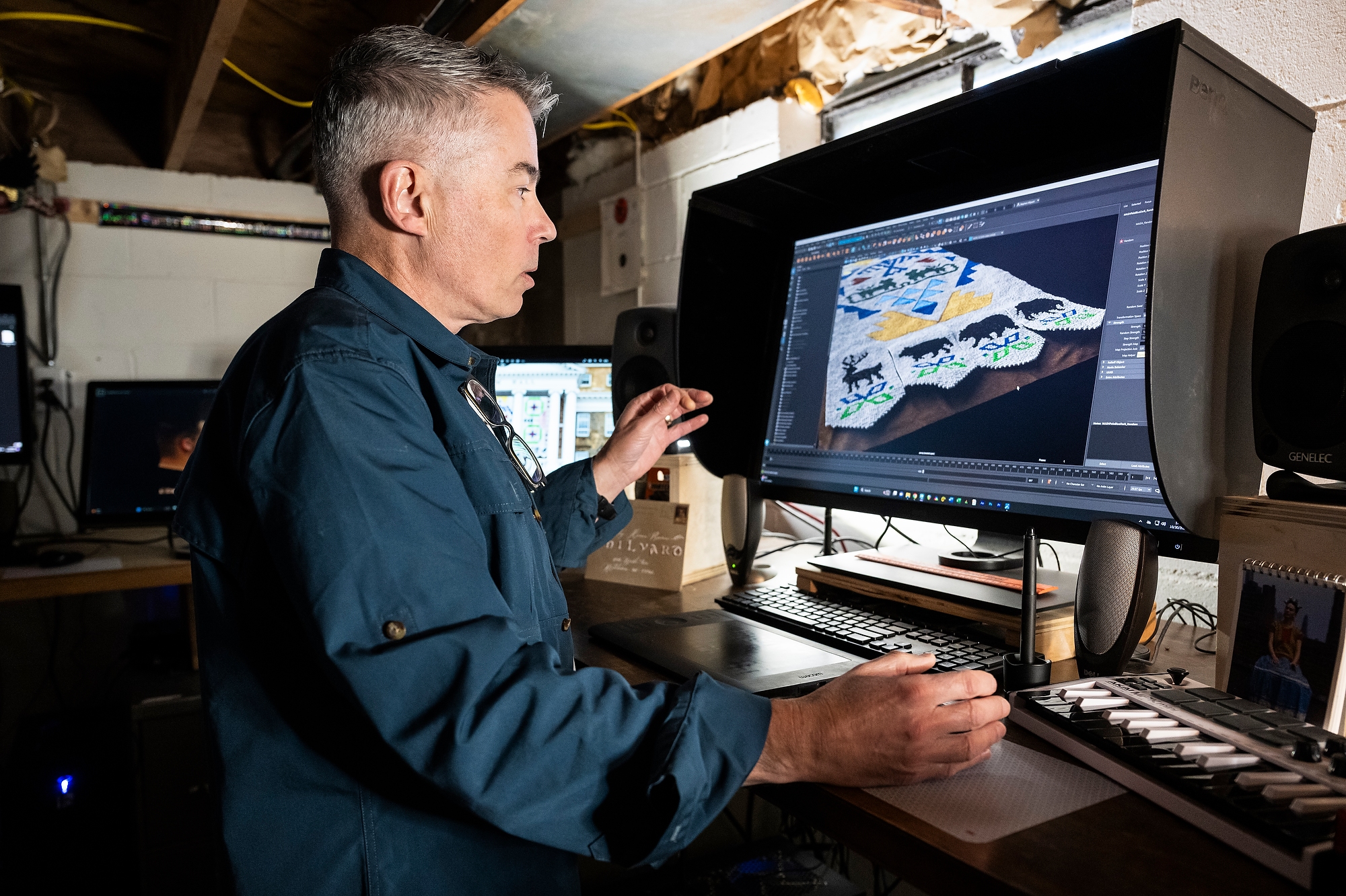 Stephen Hilyard stands at a computer monitor and speaks as he talks about the computer-generated image of the Seed by Seed banners shown on the screen. He is in his home studio, a basement space with open rafters. Most of the light in the image comes from the computer monitor and a small desk lamp, creating a dramatic effect.