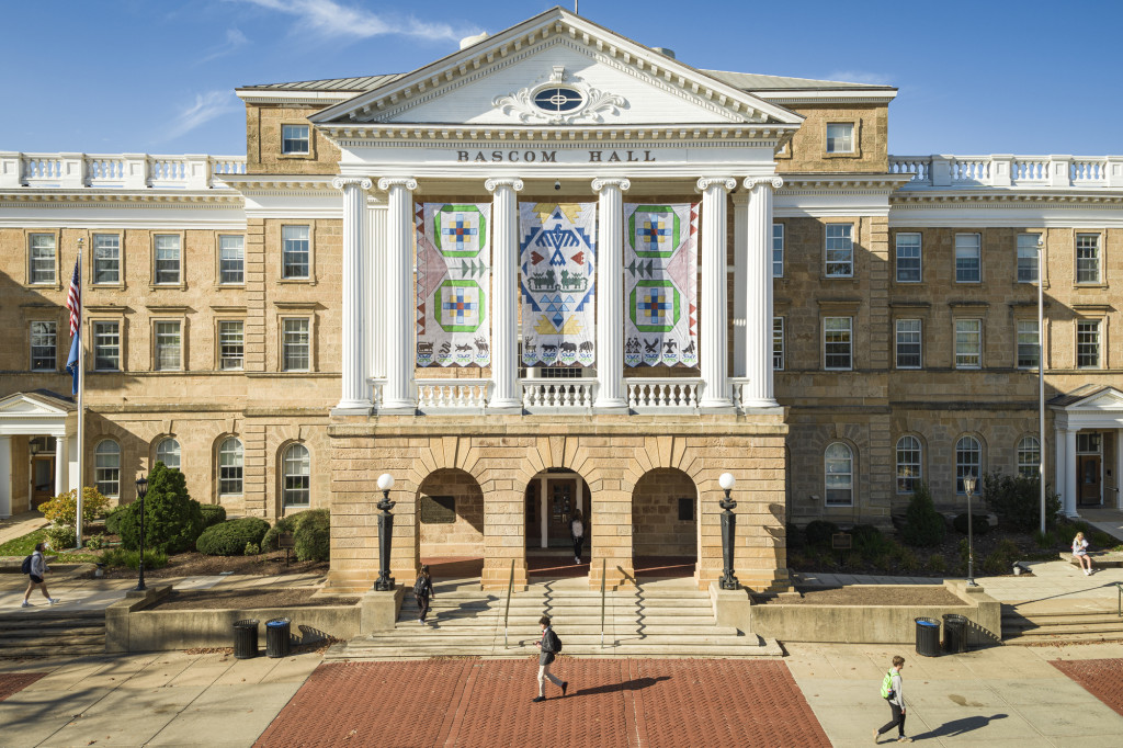 A view of the front facade of Bascom Hall taken from a drone. On a sunny day, a few people walk across the brick and concrete path in front of the building. The four panels of the Seed by Seed banner hangs between tall, white columns above the building's main entrance. The banner has been printed with a texture resembling beadwork and contains symbols and colors representing traditions of the Ho-Chunk Nation. Four green rings represent the four lakes of Teejop, the name the Ho-Chunk give the land now occupied by UW–Madison. Inside each ring, square patches in light blue, dark blue yellow and red represent the reflections of light on the water at different times of day. Two large pink triangles on either side of the banner represent flowers, with green stems and triangular leaves leading to the center panel. On the center panel, a large diamond made of small blue triangles frames a blue thunderbird, which is flanked by two red, abstract W's, representing UW–Madison. Below the thunderbird are two green water spirits, which resemble four-legged animals with very long tails. Below the water spirits are six light blue triangles representing water. Above and below the large diamond frame are bursts of yellow beading, representing the sun. Along the bottom border of the banners are stylized animal symbols of the twelve clans of the Ho-Chunk Nation, and beneath each animal is a traditional Ho-Chunk flower motif in blue and green.