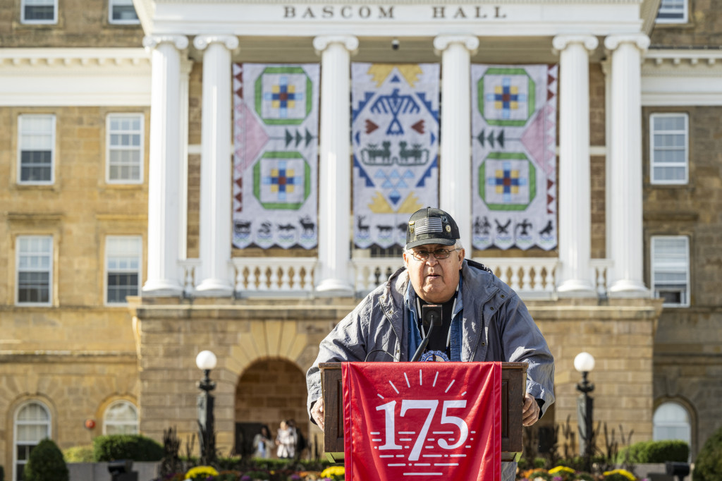 A man wearing a hat with a U.S. flag and veteran pins stands behind a podium adorned with a red cloth printed in white characters with the number 175. He speakes into a microphone. Behind him is Bascom Hall, where the Ho-Chunk banners are on display.