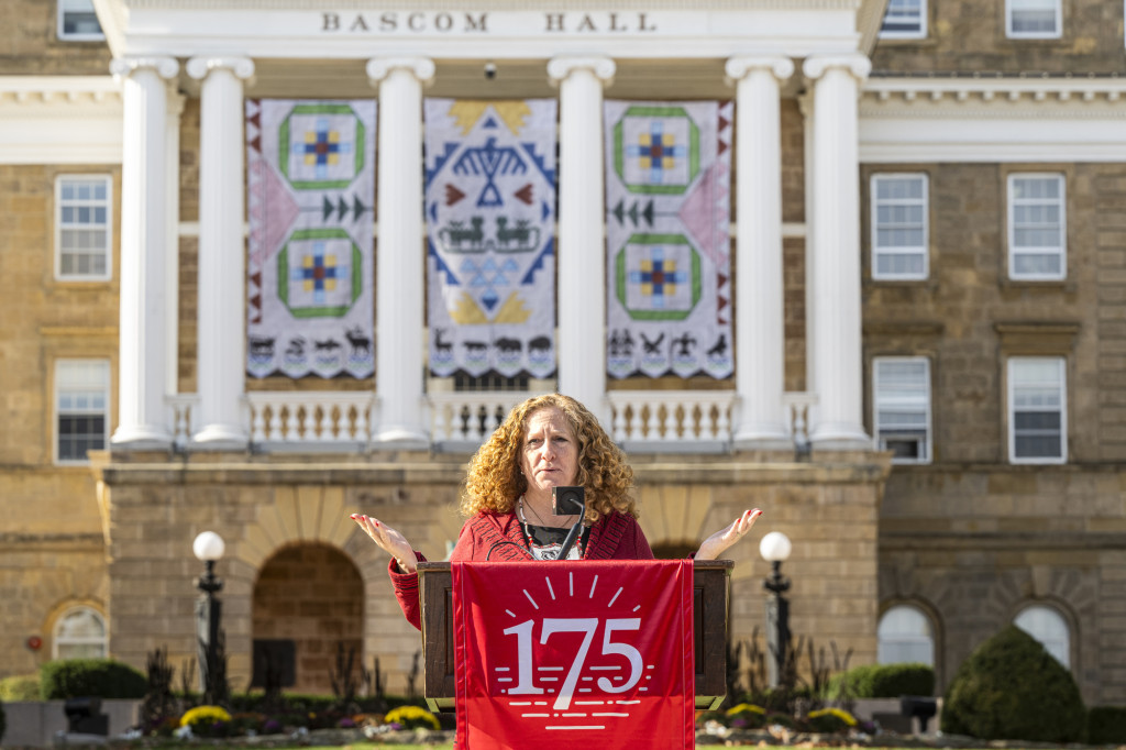 Chancellor Mnookin stands behind a podium with a red banner hanging from the front with the number 175 printed in white text. She is speaking into a microphone and gesturing with her hands. Behind her is Bascom Hall with the Ho-Chunk banners displayed between the building's columns.