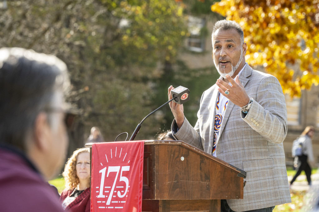 A man wearing a light brown sports jacket jestures with his hands as he speaks from the podium.