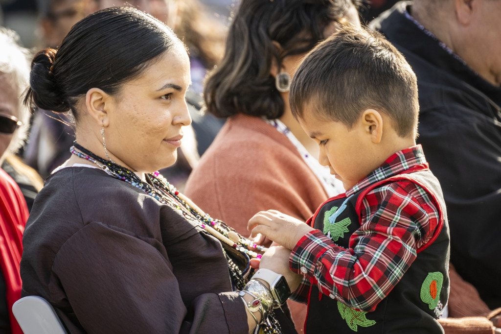 A woman seated in the audience looks on as she holds her young son, who is playing with the beaded necklaces around her neck.