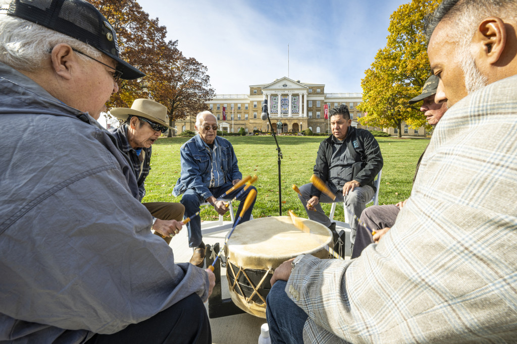 Six men seated in a circle beat a large drum with traditional mallets and sing. Behind them is Bascom Hall, decorated with the Ho-Chunk Banners.