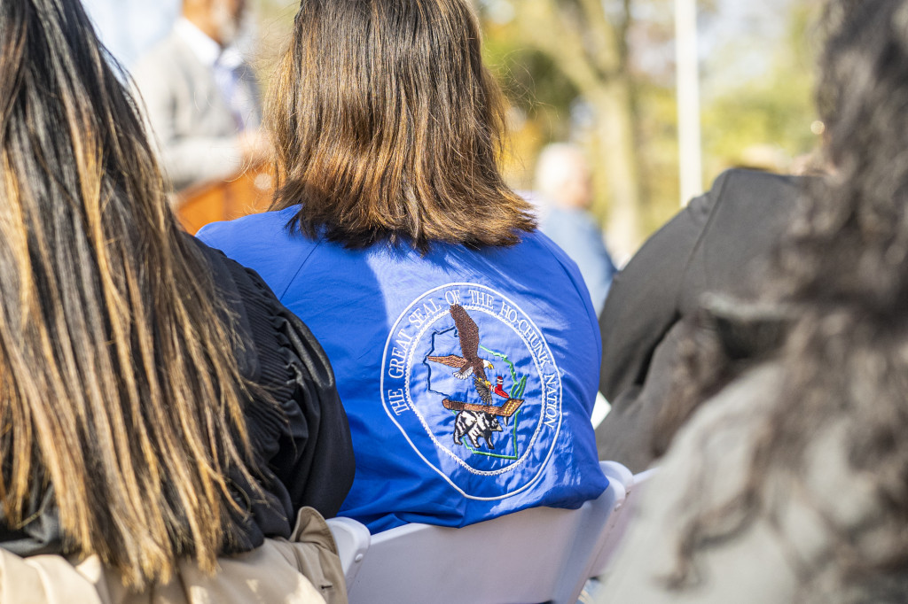 A close-up of the seated audience. One person in the audience is wearing a blue jacket with the great seal of the Ho-Chunk Nation embroidered on the back.