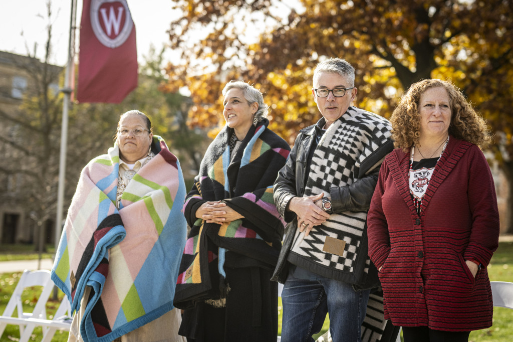 Pauliot, Fairbanks, Hilyard and Mnooking stand in a row on Bascom Hill. Pauliot, Fairbanks and Hilyard are wearing wool blankets presented to the artists on behalf of the university in thanks for their work. Mnookin is wearing a beaded pendant with an emblem of Bucky Badger, a gift to her from the Ho-Chunk Nation.