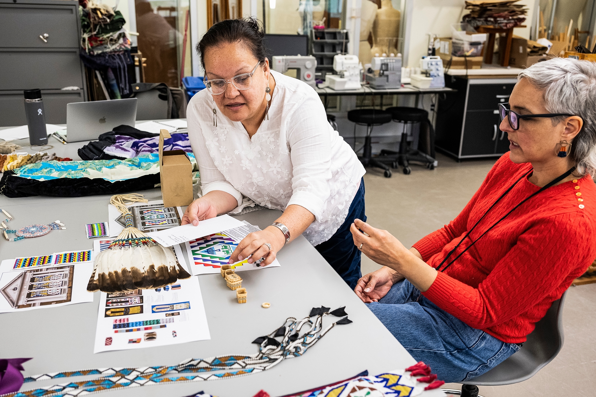 Two women speak to each other as they handle and point to objects and printed images on a large work surface in a textile design studio. The table is covered in articles of Ho-Chunk beadwork, small woven baskets and printed images of the front of Bascom Hall.