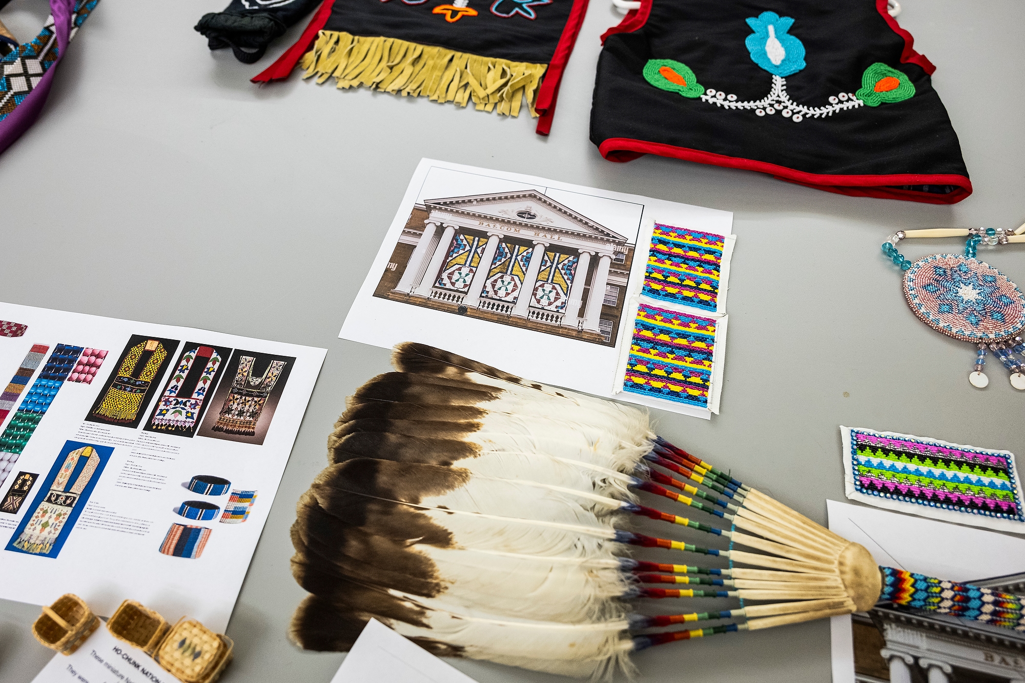 An overhead view of the table where Pauliot and Fairbanks are working. There are small woven baskets, printed images of beaded bags, a printed mockup of banners on Bascom Hall and pieces of beaded Ho-Chunk regalia, including a feather fan, garments, and bags.