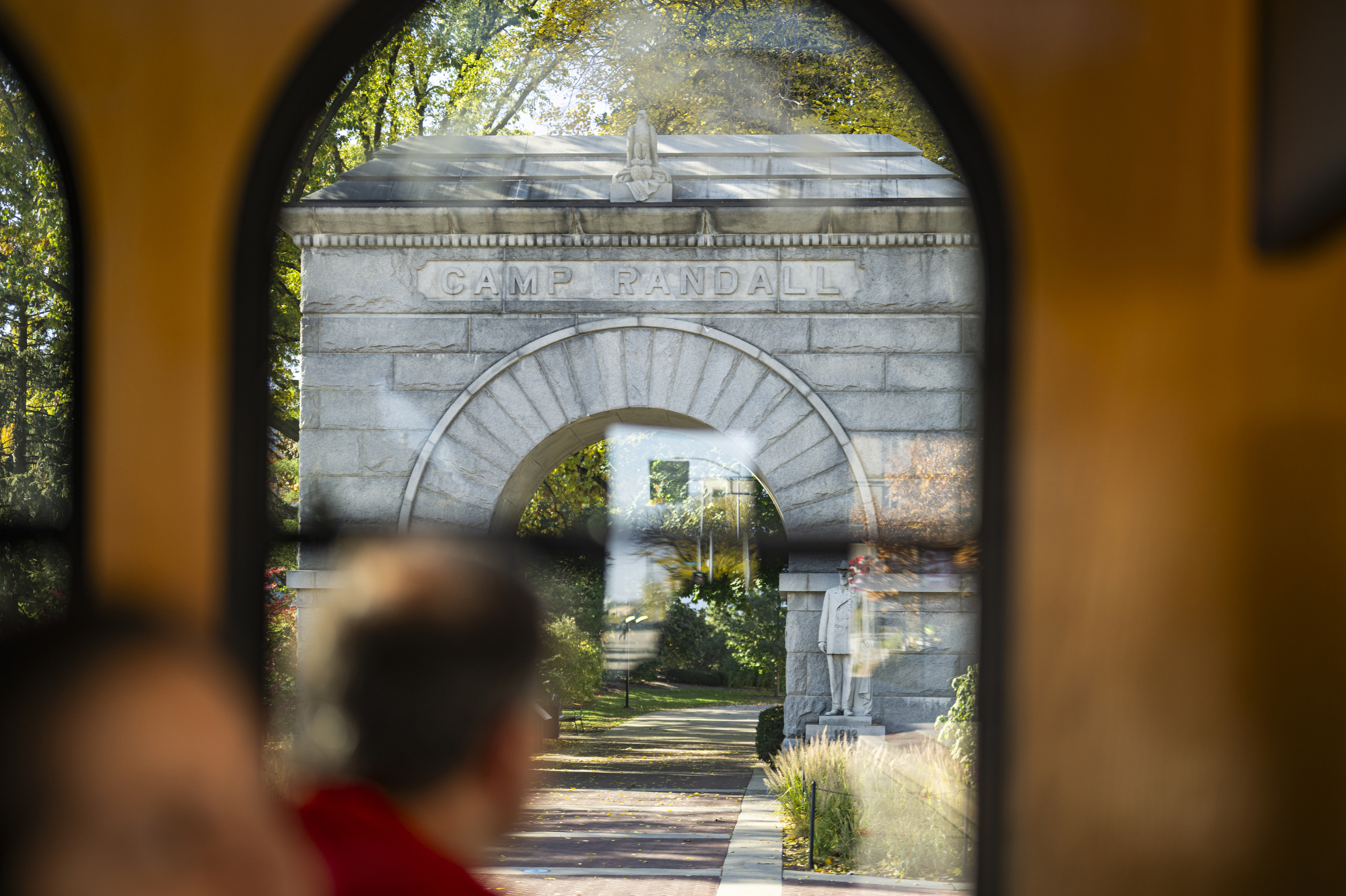 Through the window of a trolly-style bus, a passenger looks out at a stone archway with the words Camp Randall inscribed across the top.