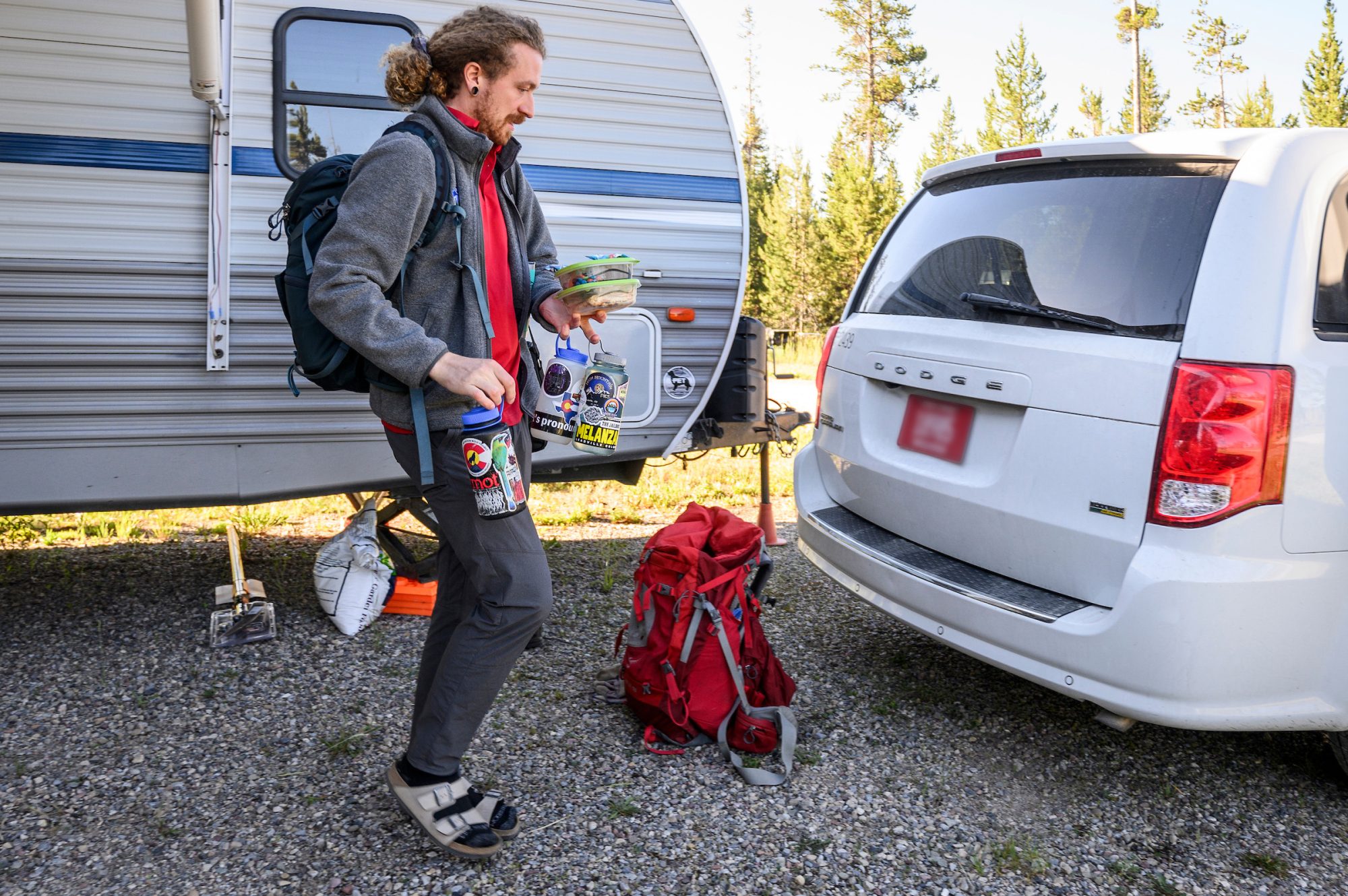 Timon Keller balances stacked containers holding his lunch and three large water bottles hanging from his fingers as he walks from the large RV to the white mini van the researchers use to drive around the parks.