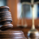 A wooden gavel sits on a wooden surface in the foreground. Blurred in the background is a brass scale, symbolizing justice.