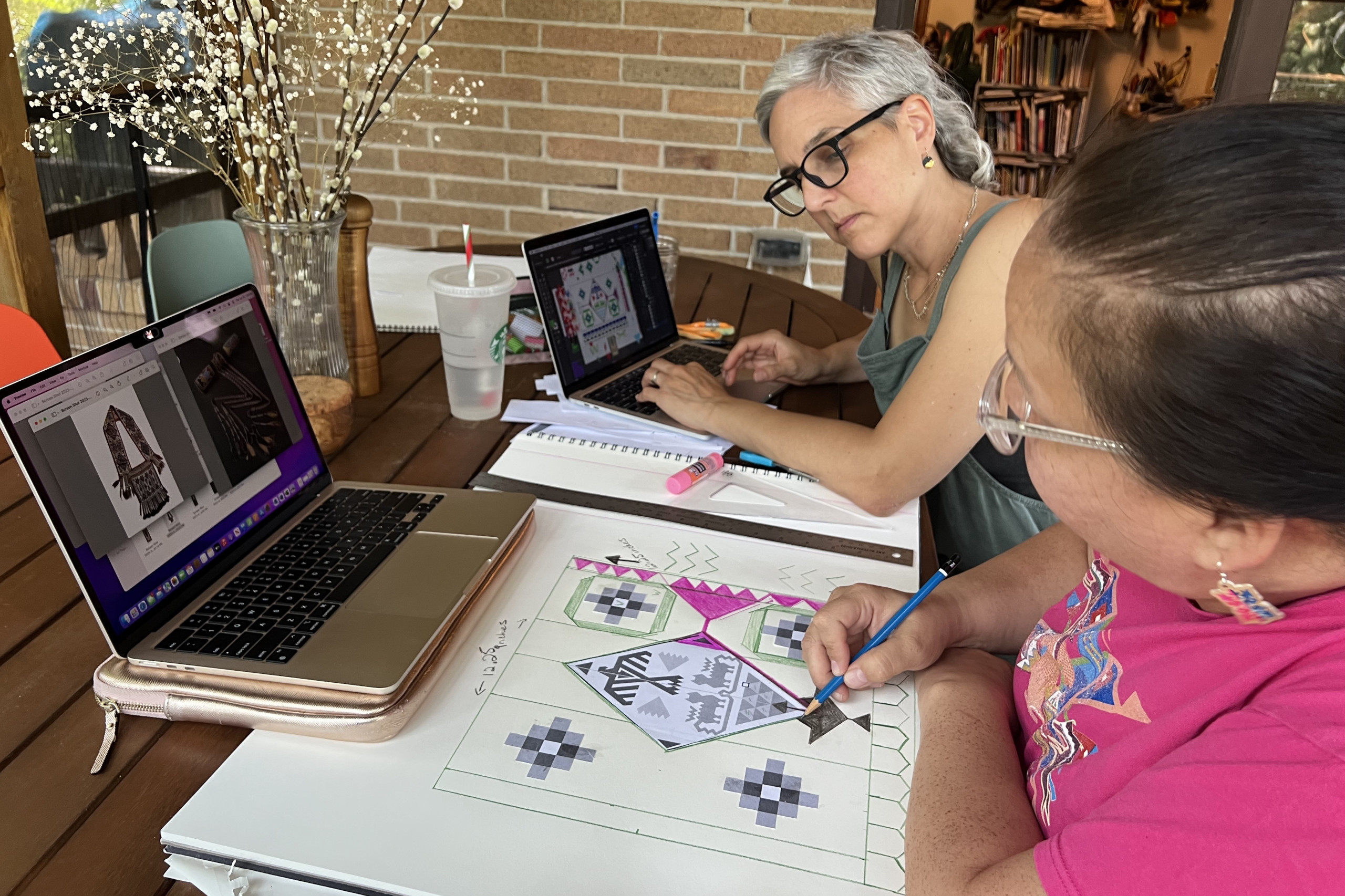 Sitting at a table on a screened porch, Pauliot draws with colored pencils in a large sketchbook. Fairbanks sits beside her. They both have laptops open to images of traditional beadwork.