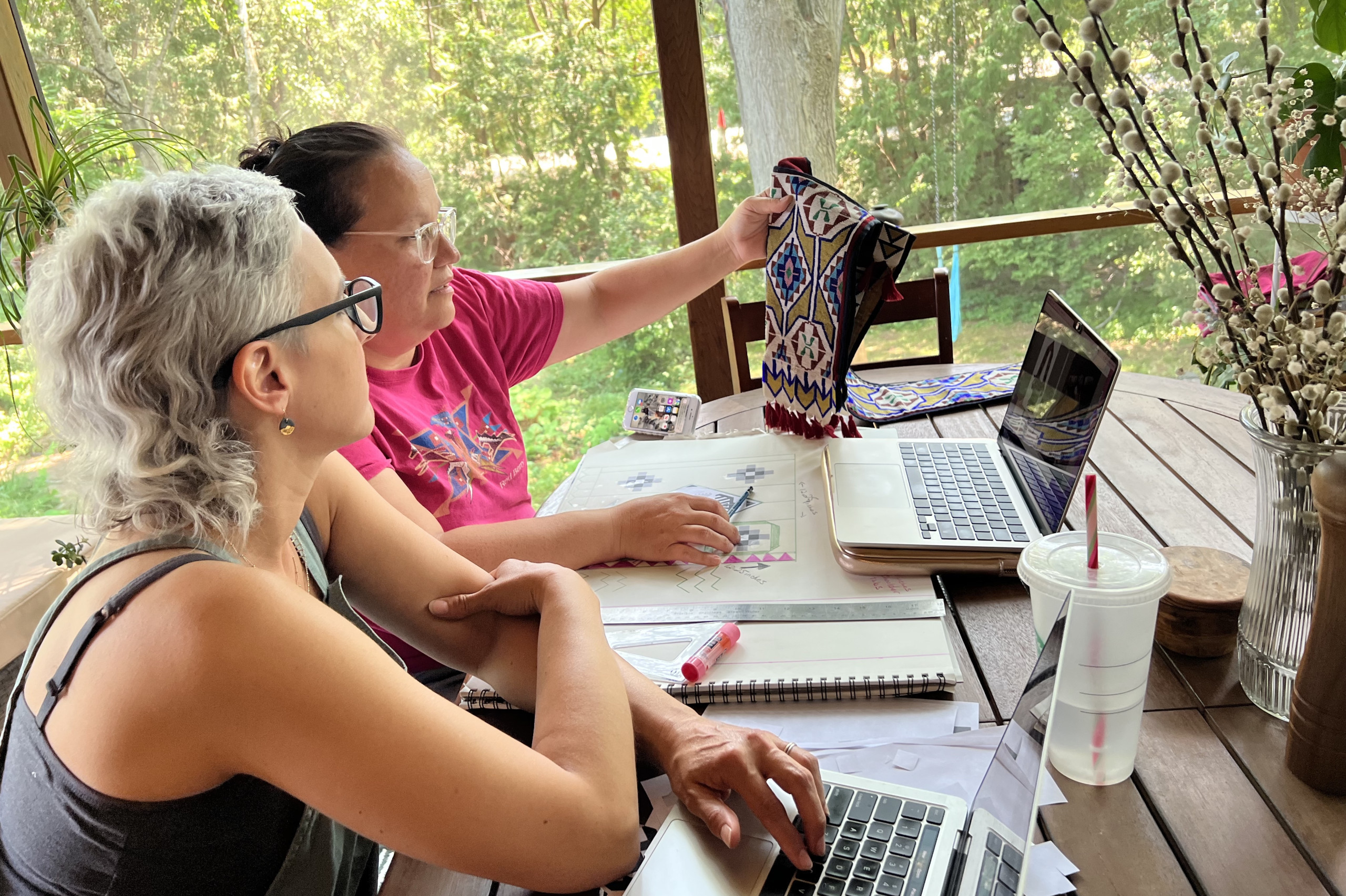 Sitting at a wooden table on a screened porch, Pauliot holds up an intricately beaded bag for Fairbanks to see.