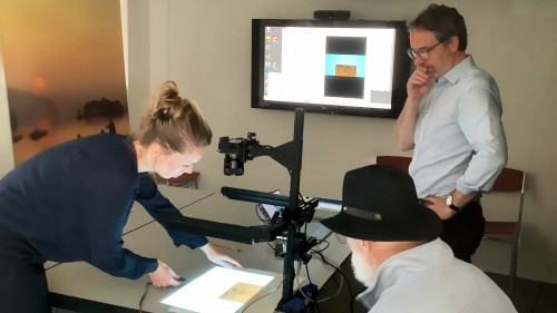 Three people work at a table. They're using a table-mounted scanning device to capture an image of an old piece of paper. On a wall-mounted monitor, there's an image of the piece of paper being captured by the scanner.