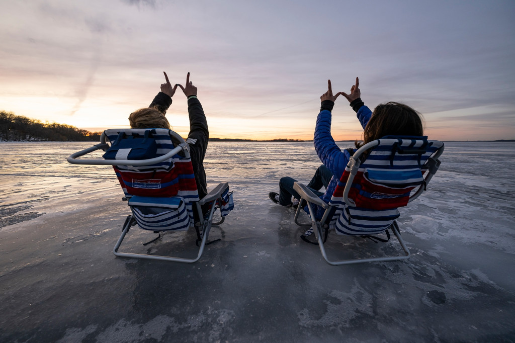Two people sit in lawn chairs with their backs to the camera. They're sitting on the ice of frozen lake Mendota and watching the sun set. They each raise their hands overhead to make a W sign with their thumbs and forefingers.