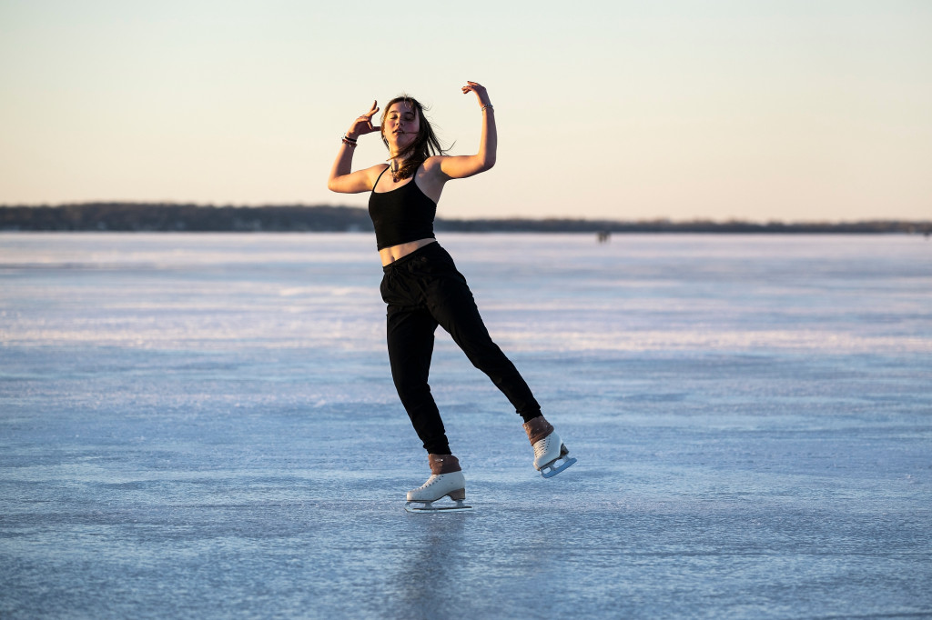 A woman wearing a black tank top and black sweatpants and white ice skates glides across the Lake Mendota ice with her arms stretched graceful overhead and her eyes closed.